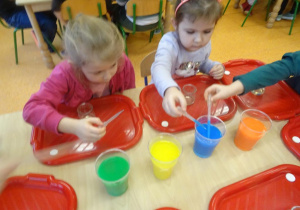 Dwoje dzieci nabiera zabarwioną ciesz stojącą w pojemnikach na środku stolika.